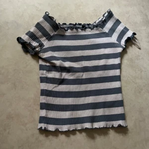 Randig ribbad topp från Blue Seven - Säljer en ribbad topp från Blue Seven med grå och vita horisontella ränder. Toppen har korta ärmar, volangkant vid halsringning och ärmslut samt en croppad passform. Perfekt för en avslappnad stil.