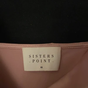 Ljusrosa långklänning från Sisters Point - Säljer en elegant ljusrosa långklänning från Sisters Point. Klänningen har smala axelband, omlott framtill och en lätt glansig satinliknande yta. Perfekt för speciella tillfällen och har en slits bak för extra rörelsefrihet.