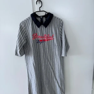 Randig klänning med krage från Emily Casual - Grå kortklänning med svarta vertikala ränder och mörkblå krage. Broderad text 'Beautiful Angels' i rött och vitt framtill. Halvlånga ärmar och rak passform, perfekt för en avslappnad stil.