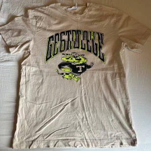 Beige t-shirt med tryck från Weekday - Säljer en beige t-shirt från Weekday med coolt tryck av en tecknad tiger och texten 'Best College' i svart och neongult. T-shirten har rund hals och korta ärmar. Perfekt för en avslappnad stil.