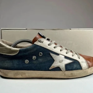 Golden Goose sneakers med stjärndetalj - Säljer ett par Golden Goose sneakers med blå jeansliknande sidor, vit stjärna och vita detaljer samt brunt skinn över tån. Skorna har snörning och GGDB-logga på plösen. Snygg och unik design med retrovibbar.