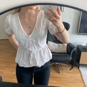 Vit blus med volang och knyt - Säljer en söt vit blus med v-ringning, puffärmar och knyt framtill. Blusen har volang nertill och är tillverkad i ett lätt bomullstyg. Perfekt för vår och sommar!