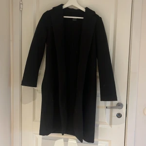 Svart lång kappa från NA-KD - Säljer en svart lång kappa från NA-KD med huva. Klassisk och stilren modell med långa ärmar och öppen framtill. Perfekt för dig som vill ha en enkel och snygg jacka till vardags.
