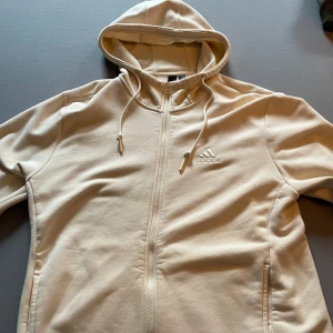 Beige hoodie från Adidas - Säljer en beige hoodiejacka från Adidas med dragkedja framtill och justerbar huva. Jackan har två fickor på sidorna och Adidas-logga broderad på bröstet. Perfekt för en avslappnad stil och enkel att matcha med det mesta. Hoodien är använd HEMMA ca 1,2 gånger. Observera att den är stor i storleken. Storlek L.