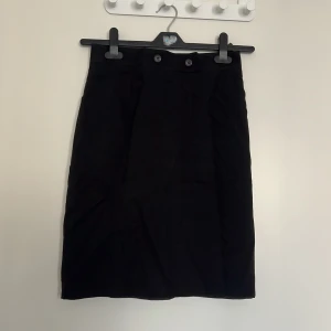 Svart pennkjol från Bonita - Klassisk svart pennkjol från Bonita med hög midja och två dekorativa knappar framtill. Kjolen är tillverkad i polyester och har dragkedja bak. Perfekt för en stilren och tidlös look.