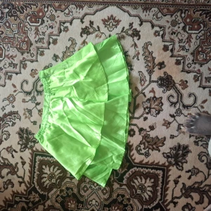 Grön volangkjol - Säljer en neonljusgrön kort kjol med volanger. Kjolen har resår i midjan och är tillverkad i ett glansigt syntetmaterial. Perfekt för dig som vill sticka ut med en färgstark look.