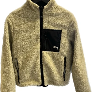 Beige och svart teddyjacka från Stussy - Teddyjacka från Stussy i beige och svart med dragkedja framtill, svart bröstficka med logga och två sidofickor. Baksidan har en stor svart panel med en vit 8-boll. Jackan har hög krage och är fodrad för extra värme.