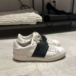 Valentino Open sneakers med svart band - Säljer ett par vita Valentino Open sneakers med ett brett svart band över mitten. Skorna har snörning, rund tå och platt sula. Klassisk och stilren design med subtila nitar på hälen.