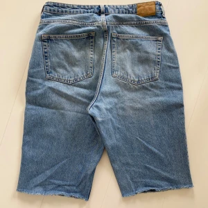 Ljusblå jeansshorts med rå kant - Säljer ett par ljusblå jeansshorts med klassisk femficksdesign och rå, avklippt kant nertill. Shortsen har normal passform och är tillverkade i denim. Perfekta för sommaren och ger en avslappnad look. Midjemått 42cm, benlängd innerlår 34cm. Kan klippas ned mera efter önskad längd.