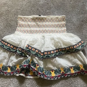 Broderad vit kortkjol från Zara - Superfin vit kortkjol från Zara med broderade blommor i gult, rött och grönt. Kjolen har smock i midjan, volanger och dekorativt knytband med tofsar. Perfekt för dig som gillar färgglada detaljer och boho-stil. 