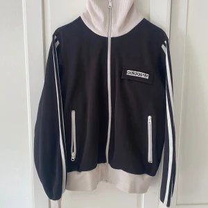 Svart och vit Adidas track jacket - Säljer en svart och vit Adidas track jacket med dragkedja framtill och två fickor med dragkedja. Jackan har klassiska vita ränder längs ärmarna och hög krage i kontrastfärg. Adidas-logga på bröstet. Strl 42 men skulle säga strl M-L