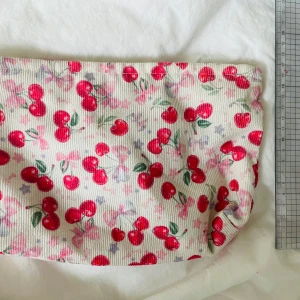 Necessär med körsbärsmönster - Söt necessär med vitt tyg och tryck av röda körsbär och rosa stjärnor. Väskan har dragkedja upptill och är perfekt för smink eller småprylar. Materialet är ribbat tyg, troligen bomull. Storlek ca 23x16 cm.