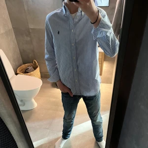 Blåvit randig skjorta från Ralph Lauren - Säljer en klassisk blå och vit randig skjorta från Ralph Lauren med button-down krage och broderad logga på bröstet. Skjortan har långa ärmar och vita knappar framtill. Perfekt för en stilren look.