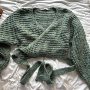 Grön stickad omlottkofta från Zara - Mysig grön stickad omlottkofta från Zara med knytband i midjan och v-ringad hals. Koftan har långa ärmar och en ribbad struktur som ger en trendig look. Perfekt att slänga på sig över en topp eller klänning.