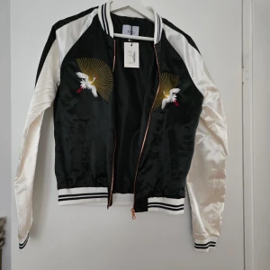 Svart och vit tunn jacka med broderad trana - Säljer en svart och vit bomberjacka från Lager 157 i blank polyester. Jackan har broderade tranor med guldstrålar på bröstet, ribbade muddar och dragkedja framtill. Perfekt statement-plagg för dig som vill sticka ut. Katt finns I hemmet
