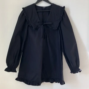 Svart klänning med sjömanskrage från Monki - Svart klänning från Monki med sjömanskrage och knytband framtill. Långa ärmar med resår vid ärmslut och volangdetaljer längs krage och fåll. Perfekt för en trendig look. Endast använd en gång. 