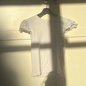 Vit topp med spetsärmar från Gina Tricot - Säljer en söt vit topp från Gina Tricot med ärmar i spets och broderade blommor. Toppen har rund halsringning och är i mjukt material. Perfekt för en fräsch och somrig look!