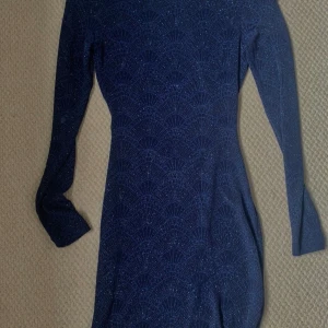 Mörkblå glittrig långärmad klänning - Säljer en mörkblå glittrig klänning med lång ärm och snygg öppen rygg. Klänningen har ett diskret mönster och en tight passform som framhäver figuren. Perfekt för dig som vill sticka ut lite extra!