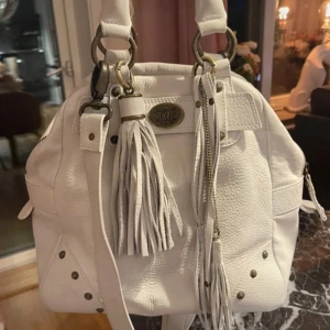 Vit handväska med tofsdetaljer - Säljer en snygg vit handväska från SRL med stora tofsar framtill och dekorativa nitar i hörnen. Väskan har dubbla handtag och en metallbricka med logga. Perfekt för dig som vill ha en stilren accessoar med lite edge.