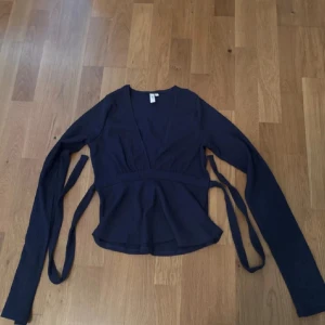 Mörkblå omlottopp från Nelly - Säljer en mörkblå omlottopp från Nelly med långa ärmar och knytband i midjan. Toppen har v-ringning och är tillverkad i ett mjukt, ribbat material. Perfekt för att skapa en snygg och feminin siluett.