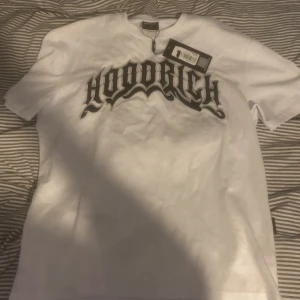 Vit t-shirt från Hoodrich med svart tryck - Säljer en vit t-shirt från Hoodrich i storlek XS. HELT NY, T-shirten har ett stort svart tryck med Hoodrich-loggan på bröstet. Klassisk passform och rund halsringning. Perfekt för dig som gillar streetwear. 