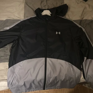 Svart och grå vindjacka från Under Armour - Säljer en snygg svart och grå vindjacka från Under Armour med huva och dragkedja framtill. Jackan har lång ärm och logga på bröstet. Perfekt för utomhusaktiviteter och träning.