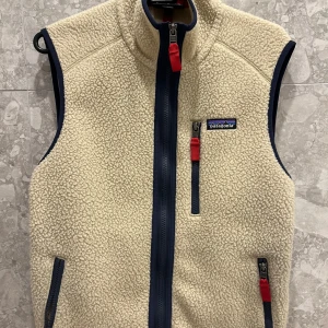 Beige fleeceväst från Patagonia - Säljer en beige fleeceväst från Patagonia i storlek S med marinblå detaljer och röda dragkedjor. Västen har hög krage, tre fickor med dragkedja och klassisk Patagonia-logga på bröstet. Perfekt för lager-på-lager och chill utomhus. 