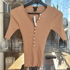 Beige ribbad topp från Lindex - Snygg beige ribbad topp från Lindex med v-ringning och dekorativa knappar framtill. Modellen har halvlånga ärmar och en figurnära passform. Prislappen sitter kvar, nypris 400kr