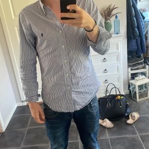 Randig skjorta från Ralph Lauren - Säljer en klassisk skjorta från Ralph Lauren med smala blå och vita ränder. Skjortan har lång ärm, knappar framtill och ikoniska broderade loggan på bröstet. Tillverkad i bomull och har en normal passform. Perfekt för dig som gillar stilren och tidlös look.