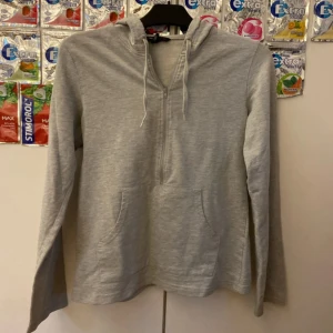 Ljusgrå hoodie med half zip och huva - Ljusgrå hoodie från H&M med huva och snörning, half zip-dragkedja framtill och känguruficka. Mjuk bomullskänsla och broderad H&M-logga på fickan. Perfekt för chill dagar eller när du vill ha en enkel och clean look.