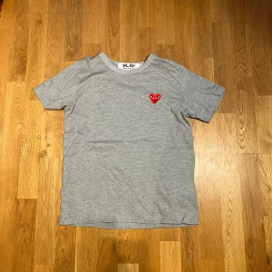 Grå t-shirt Comme des Garçons Play - Grå t-shirt från Comme des Garçons Play med klassiskt rött hjärta med ögon broderat på bröstet. T-shirten har rund halsringning och korta ärmar. Tillverkad i mjuk bomull för en skön och avslappnad vibe.