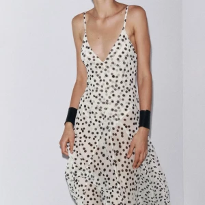 Vit lång prickig klänning med v-ringning - Supersnygg långklänning i vit, transparent tyg med svarta prickar. Djup v-ringning och tunna axelband ger en cool och trendig look. Fladdrig och luftig modell som känns perfekt för varma dagar eller när du vill sticka ut lite extra.