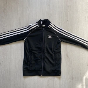 Svart Adidas kofta med vita ränder - Klassisk svart Adidas training gear kofta med vita ränder längs ärmarna och dragkedja framtill. Koftqn har ribbade muddar, ståkrage och två fickor. Ikonisk Adidas-logga på bröstet. Perfekt för en sportig och avslappnad stil.