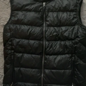 Svart dunväst med dragkedja - Säljer en svart dunväst med quiltad design och hel dragkedja framtill. Västen har två fickor med dragkedja och en klassisk, enkel look. Perfekt att slänga över en hoodie eller tröja när det är kyligt ute.