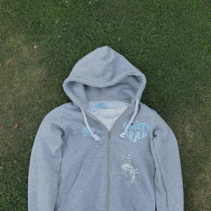 Grå tracksuit  - Säljer en grå hoodie från GRAVITY GRAB med dragkedja, huva och vita snören. Tryck i blått och vitt på bröstet och coola färgstänk som detalj. Perfekt för chill dagar eller streetwear-stil. Mjuk bomullskänsla och långärmad modell. Pris kan diskuteras vid snabb affär 