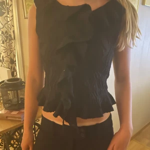 Svart volangblus från Gina Tricot - Supersnygg svart blus från Gina Tricot med volangdetaljer framtill och längs nederkanten. Toppen är ärmlös och har ett väldigt fint mönster som ger en cool och trendig look. Perfekt att ha på kalas och fester, men den går även att klä ner om du vill😽