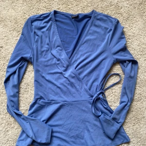 Blå omlottopp från Gina Tricot XS - Supersnygg blå omlottopp från Gina Tricot i storlek XS. Toppen har långa ärmar, v-ringning och knyt i sidan för en trendig look. Perfekt att styla med jeans eller kjol för en cool vardagsstil.