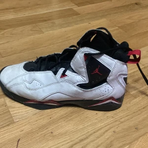 Nike Air Jordan 7 Retro svart/vit/röd - Säljer ett par Nike Air Jordan 7 Retro i svart, vitt och rött. Skorna har klassisk Jordan-design med högt skaft, snörning och coola röda detaljer. Ovandelen är i skinn och textil, med svart mesh vid plösen och röd logga på sidan. Perfekta för dig som gillar streetstyle.