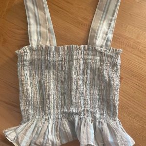 Randig linnetopp från Uniqlo XS - Supersöt randig linnetopp från Uniqlo i beige, vitt och ljusblått. Toppen har breda axelband, smockad överdel och volangkant nertill. Perfekt för varma dagar och ger en chill sommarkänsla.