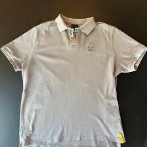 Grå pikétröja från Sail Racing M - Snygg grå pikétröja från Sail Racing i bomull. Klassisk krage med två knappar och broderad logga på bröstet. Kortärmad modell med gul detalj i nederkant. Perfekt för en clean och sportig stil.