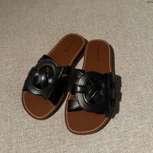 Svarta sandaler från Coach i skinn - Snygga svarta sandaler från Coach med flätad detalj över foten och platt brun sula. Tillverkade i skinn med öppen tå och stilren design. Kommer i originalkartong. Aldrig använda.