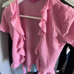 Rosa kofta med volangdetaljer - Söt rosa kofta med korta ärmar och volang längs hela kanten. Perfekt för att lägga till en feminin touch till din outfit. Lätt och mjukt material som känns skönt mot huden. Köpt från edikted aldrig använd.
