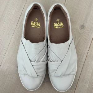 Vita slip-on sneakers från Dasia - Snygga vita slip-on sneakers från Dasia med coolt omlott-detalj över foten. Skorna har en chunky platt sula och är tillverkade i mjukt skinn för en clean och modern look.
