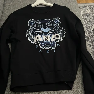 Svart Kenzo sweatshirt Strl M - Svart sweatshirt från Kenzo med ikoniskt broderat tigerhuvud i blått och vitt framtill. Tröjan har rund hals, ribbade muddar och lång ärm. Perfekt statement-plagg för dig som gillar streetstyle och snygga detaljer. Tröjan är i väldigt bra skick och knappt använd 