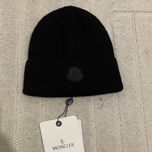 Svart ribbad mössa från Moncler - Snygg svart ribbad mössa från Moncler med broderad logotyp framtill. Perfekt för kyliga dagar och ger en stilren look. Passar både till vardags och mer uppklädda tillfällen.