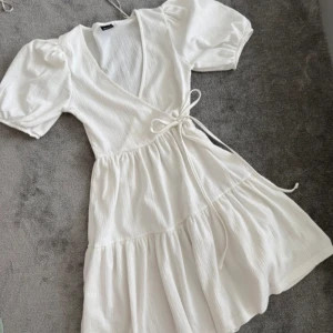 Vit omlottklänning från Gina Tricot - Supersöt vit kort omlottklänning från Gina Tricot med puffärmar och knyt i midjan. Klänningen har v-ringning och volangig kjol, perfekt för sommarens alla tillfällen. Tillverkad i ett lätt och luftigt bomullstyg.