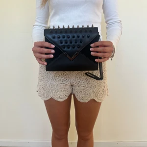 Svart kuvertväska med nitar - Cool svart kuvertväska i skinn med stora svarta nitar och spetsiga metalldetaljer på locket. Väskan har ett kuvertformat lock med metallspänne och en smal handledsrem. Perfekt för dig som vill sticka ut med en edgy accessoar.