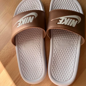 Nike sandaler i vitt och koppar - Snygga Nike sandaler med bred kopparfärgad rem och stort vitt Nike-logga. Sulan är vit med struktur för extra komfort. Perfekta för sommaren och chill dagar, riktigt fräsch färgkombo som sticker ut.