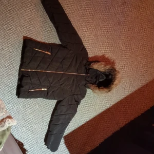 Svart barnjacka - Säljer en svart barnjacka med huva med fuskpälsdetalj. Jackan har guldiga dragkedjor på fickorna och framtill, samt ett snyggt quiltat mönster. Perfekt för kalla dagar. Huvan kan enkelt tas av och jackan bli använt utan. 