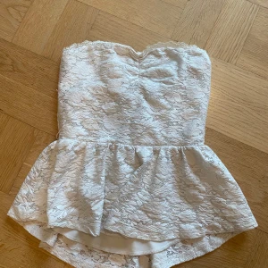 Vit spets tubtopp med volang - Superfin vit tubtopp med spets och volang nertill. Toppen är axelbandslös och har en hjärtformad urringning upptill. Fodrad under spetsen för att inte vara genomskinlig. Toppen är 31cm i överkant.